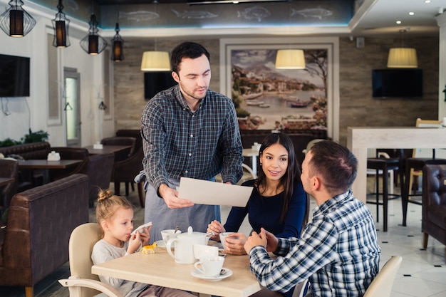 Concetto di ristorante e vacanza. cameriere dando menu alla famiglia felice al bar