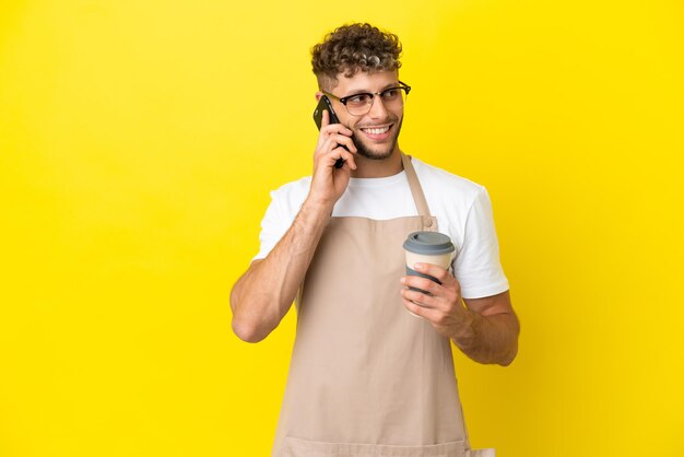 Restaurant ober blonde man geïsoleerd op gele achtergrond met koffie om mee te nemen en een mobiel