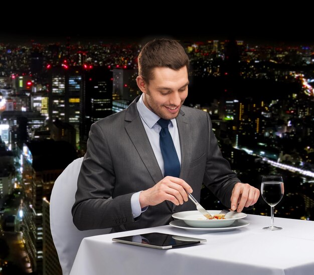 レストラン、人、技術、休日のコンセプト-レストランでメインコースを食べるタブレットPCと笑顔の男