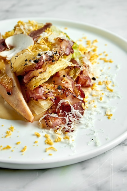 Restaurant serving of classic caesar salad with croutons, egg, mushrooms, grilled chicken, parmesan and bacon served in a white plate on a marble table