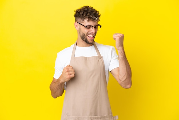 Restaurant waiter blonde man isolated on yellow background celebrating a victory