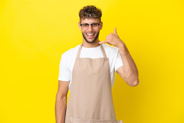 Restaurant waiter blonde man isolated on yellow background making phone gesture. Call me back sign