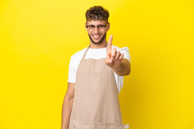 Restaurant waiter blonde man isolated on yellow background showing and lifting a finger