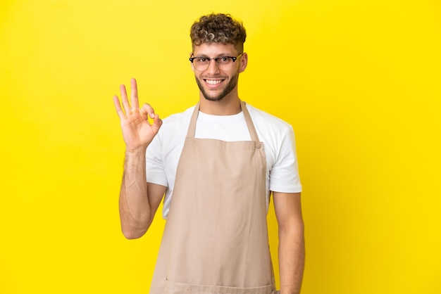 Restaurant waiter blonde man isolated on yellow background showing ok sign with fingers