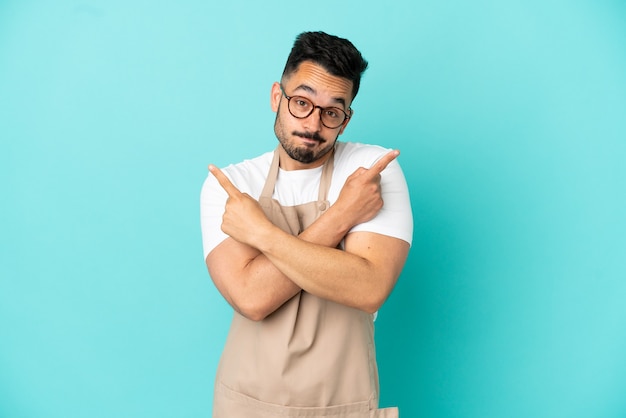 Uomo caucasico del cameriere del ristorante isolato su fondo blu che indica i laterali che hanno dubbi