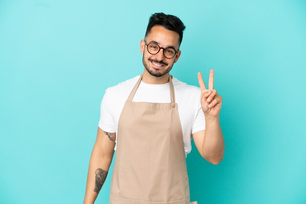 Restaurant waiter caucasian man isolated on blue background smiling and showing victory sign
