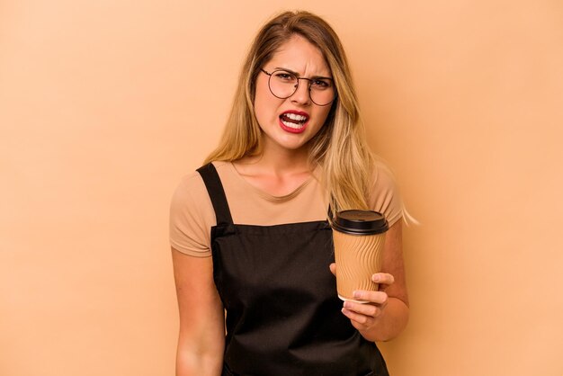 非常に怒って攻撃的な叫びベージュの背景に分離されたテイクアウトコーヒーを保持しているレストランウェイター白人女性