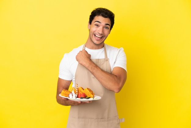Restaurant waiter holding waffles over isolated yellow background celebrating a victory