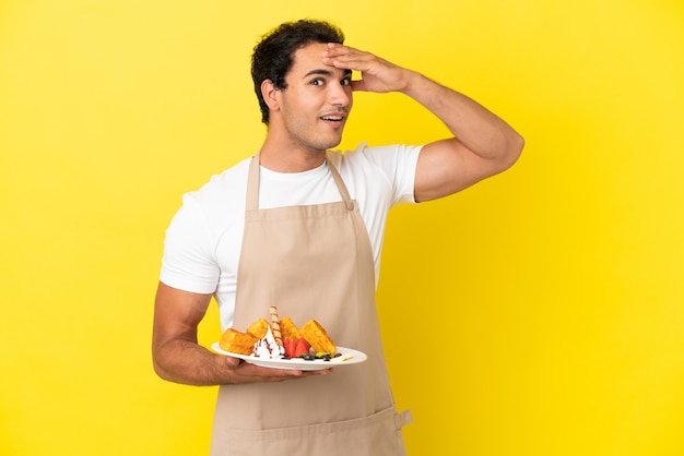 Restaurant waiter holding waffles over isolated yellow background doing surprise gesture while looking to the side