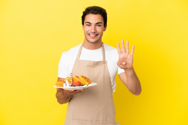 Официант ресторана держит вафли на изолированном желтом фоне счастливым и считает четыре пальцами