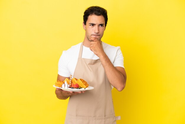 Restaurant waiter holding waffles over isolated yellow background having doubts and thinking