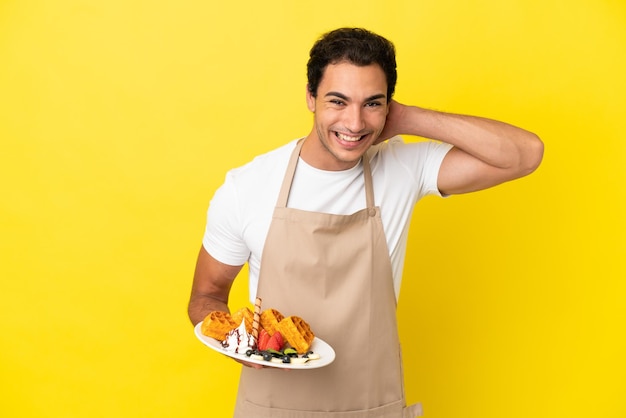 Restaurant waiter holding waffles over isolated yellow background laughing