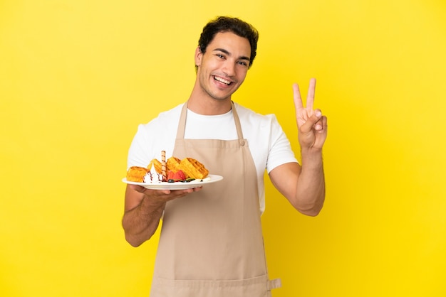 Официант ресторана держит вафли на изолированном желтом фоне, улыбаясь и показывая знак победы