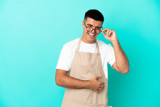 Restaurant waiter man over isolated blue background with glasses and happy