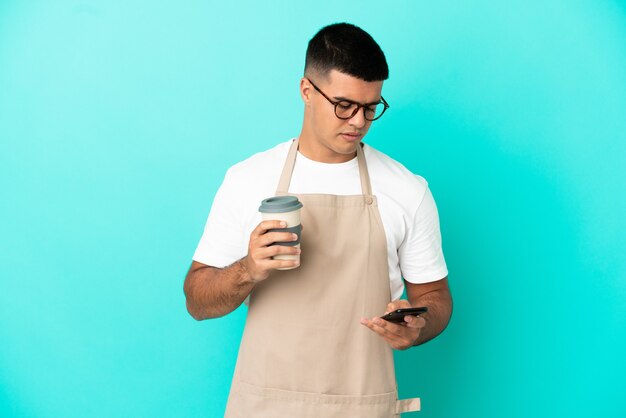 Restaurant waiter man over isolated blue wall holding coffee to take away and a mobile