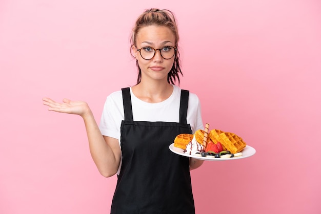 Restaurant waiter Russian girl holding waffles isolated on pink background having doubts while raising hands