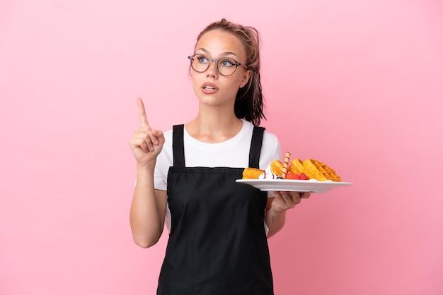 Restaurant waiter Russian girl holding waffles isolated on pink background thinking an idea pointing the finger up