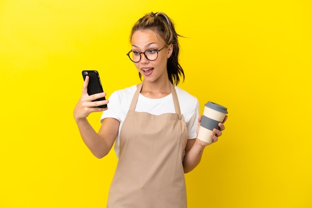 Русская девушка официант ресторана изолирована на желтом фоне держит кофе на вынос и мобильный