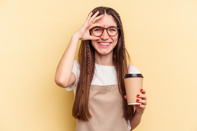 黄色の背景に分離されたテイクアウトコーヒーを持っているレストランのウェイターの女性は、目で大丈夫なジェスチャーを維持して興奮しています。