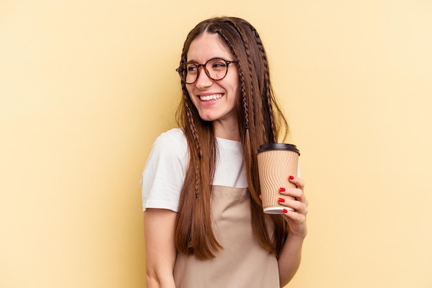 Restaurant waiter woman holding a take away coffee isolated on yellow background looks aside smiling cheerful and pleasant