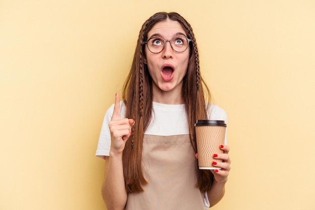 Restaurant waiter woman holding a take away coffee isolated on yellow background pointing upside with opened mouth.