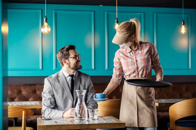 Restaurant work and corona virus. Waitress is wearing a uniform and a protective face mask. She leaves a bottle of water and a cup of coffee on the table and serves the guest in a suit in a restaurant