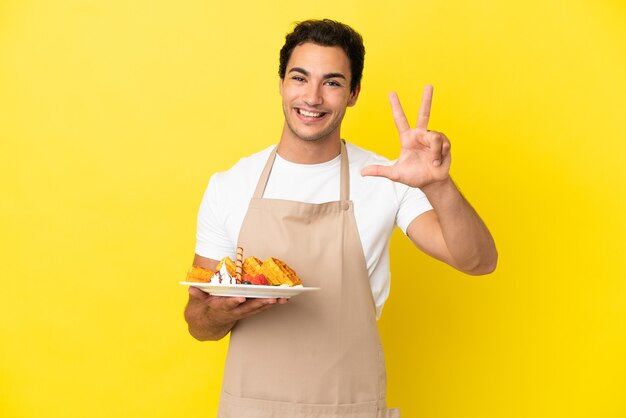 Restaurantkelner die wafels over geïsoleerde gele achtergrond gelukkig houdt en drie met vingers telt