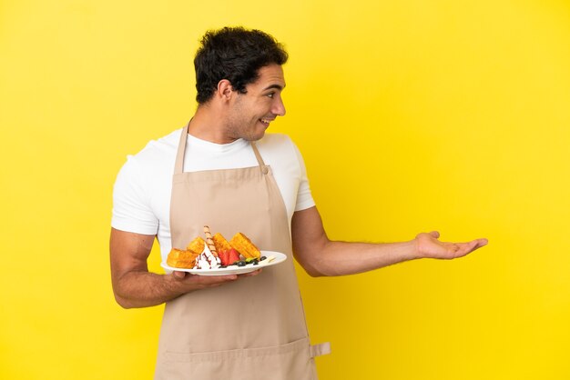 Restaurantkelner die wafels vasthoudt over geïsoleerde gele achtergrond met verrassingsuitdrukking terwijl hij opzij kijkt