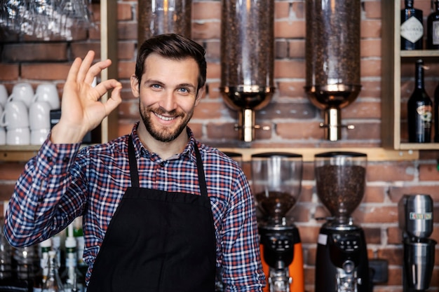 Restaurantservice en café. Een volwassen mannelijke ober staat achter een bar in een café en toont een handteken van goedkeuring. Een aangename glimlach en een positieve instelling. Cafetaria en koffiezetten