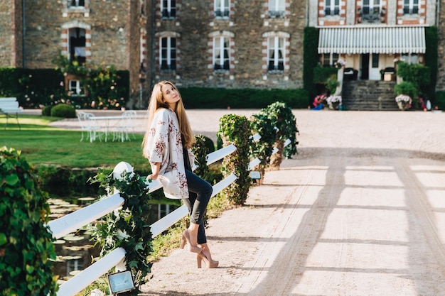 Restful female spends her free time walks near old ancient brick castle leans at white hence wears fashionable clothes and high heeled shoes enjoys sunny day and beautiful exterior outdoor