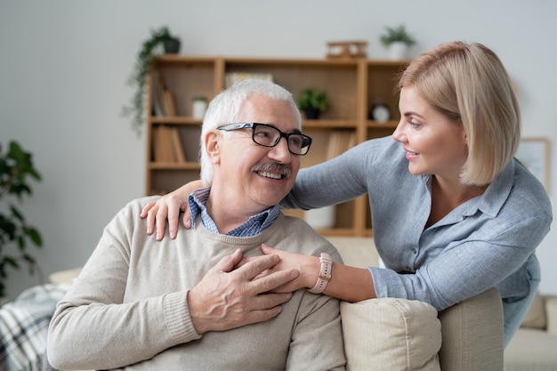 Restful senior man on couch looking at his young blonde daughter standing near by, smiling at her father and embracing him