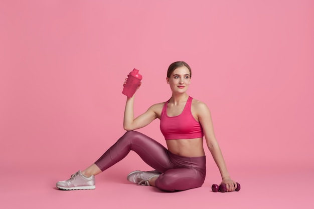 Resting. Beautiful young female athlete practicing , monochrome pink portrait. Sportive fit caucasian model with drinking bottle. Body building, healthy lifestyle, beauty and action concept.