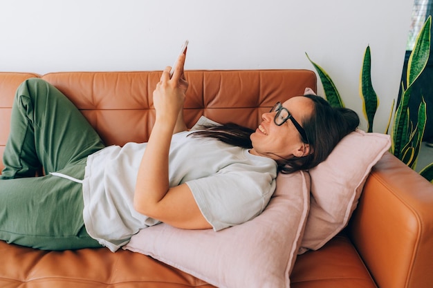 Resting laughing brunette lying on the sofa uses a mobile phone to chat