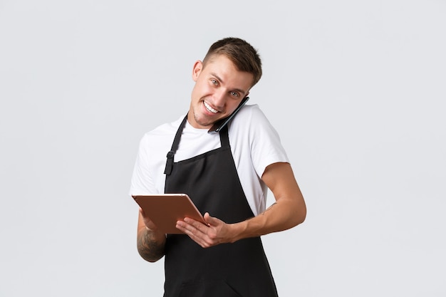 Retail stores, small business, cafe and restaurants takeaway concept. Handsome young shop manager, employee receiving order over phone, talking with customer and writing down info in digital tablet