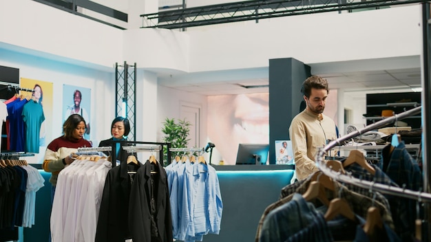 Retail worker helping customer to buy shirts at mall, diverse people looking at new fashion collections. Male client and store assistant examining trendy clothes, commercial activity.