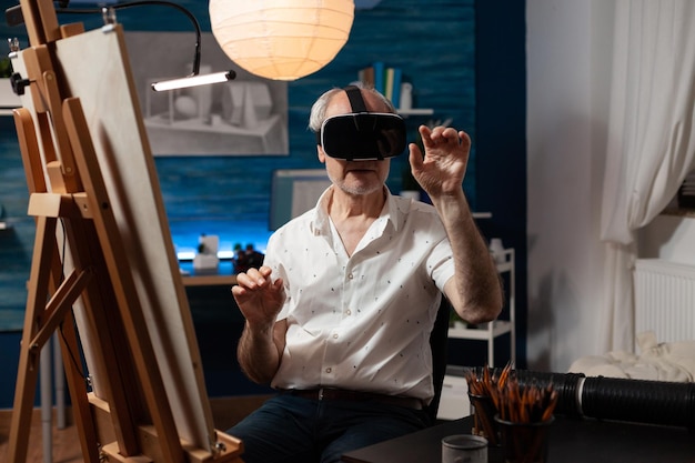 Retired art teacher with modern technology vr goggles looking at 3d artwork in creative workshop. Senior sketch artist using virtual reality glasses doing air gesture improving freehand technique.