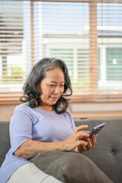 A retired Asian woman using her phone on a sofa in the living room chatting scrolling on phone