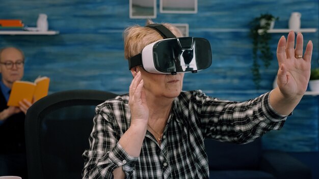 Retired concentrated senior woman using virtual reality goggles in living room gesturing sitting in home workplace. Senior woman experiencing vr headset while man sitting on sofa and reading a book