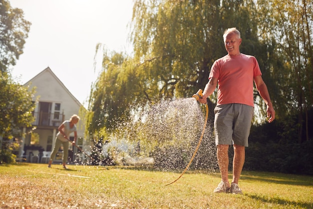 Retired Couple At Work Watering Plants With Hose And Tidying Garden With Rake At Home