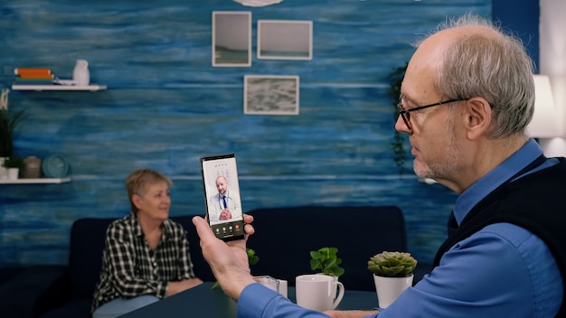 Retired male holding smartphone during online consultation listening young physician doctor sitting ...