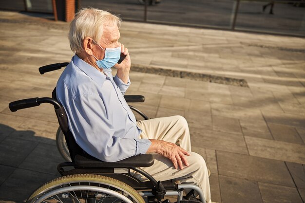 Retired man having a walk in a wheelchair near a glass building while talking on mobile phone in the city