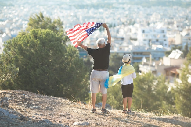 Retired man and kid boy holding the flags of the USA and Ukraine on city view