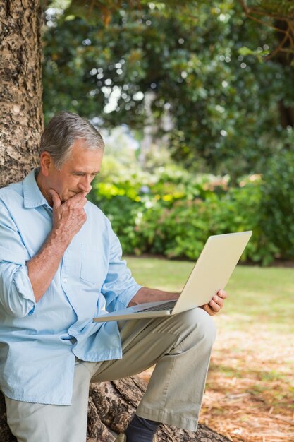 Retired man leaning against tree with a laptop