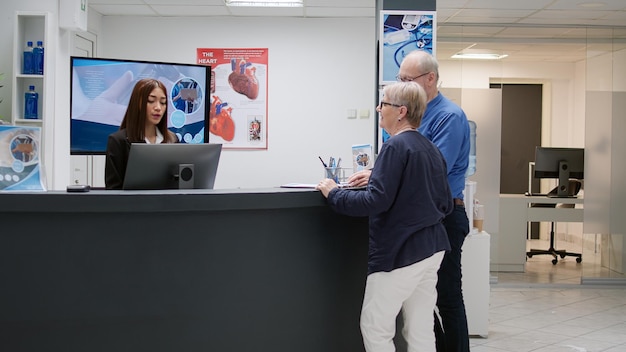 Retired people doing medical report with receptionist in hospital waiting room, woman at reception desk helping patients with checkup appointment. Assistant working in healthcare center.