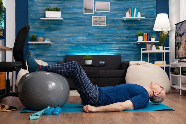 Retired senior sportman sitting on yoga mat practicing warming legs up exercises using swiss ball stretching abdominals muscles
