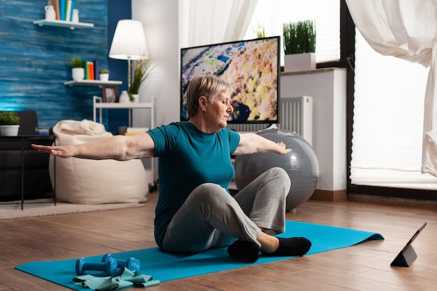 Retired senior woman searching fitness tutorial on laptop sitting on yoga mat streching arm during w...