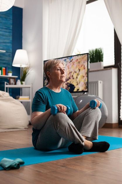 Retired senior woman sitting on yoga mat holding fitness dumbbells in lotus position during pilates wellness routine