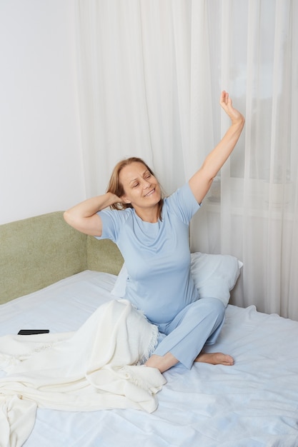 Retired woman in blue pajamas stretching in the morning after sleeping in her bed.