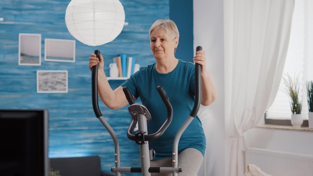 Retired woman training with stationary bicycle at home. Senior adult doing workout and physical exercise while using cardio bike machine for wellness and endurance. Pensioner cycling