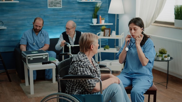 Retired woman in wheelchair receiving consultation from nurse. Medical assistant using stethoscope for healthcare checkup in nursing home. Patient with disability getting assistance.